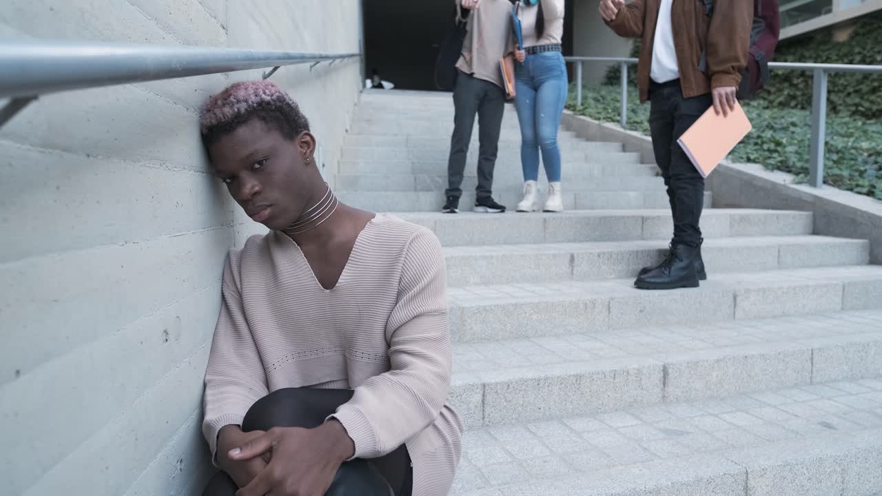 Sad african transgender person sitting while a group of students pointing to him