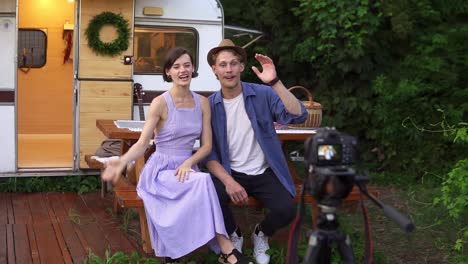 Popular-Bloggers-Couple-Greeting-Their-Followers,-Recording-Video-While-Sitting-At-The-Wooden-Table-In-Front-The-Wheels-House,-Talking-And-Looking-At-Camera-On-Tripod