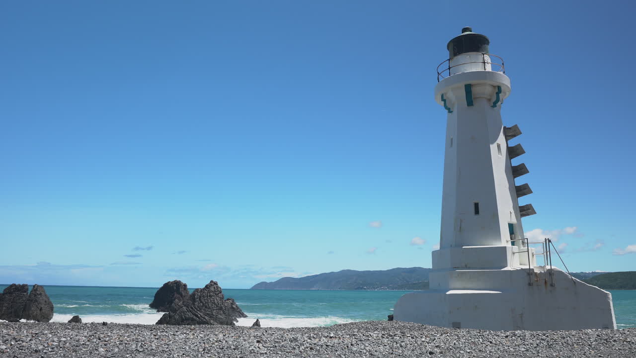 A Female cyclist rides past Pencarrow Lighthouse from left to right on a sunny day on an E-bike
