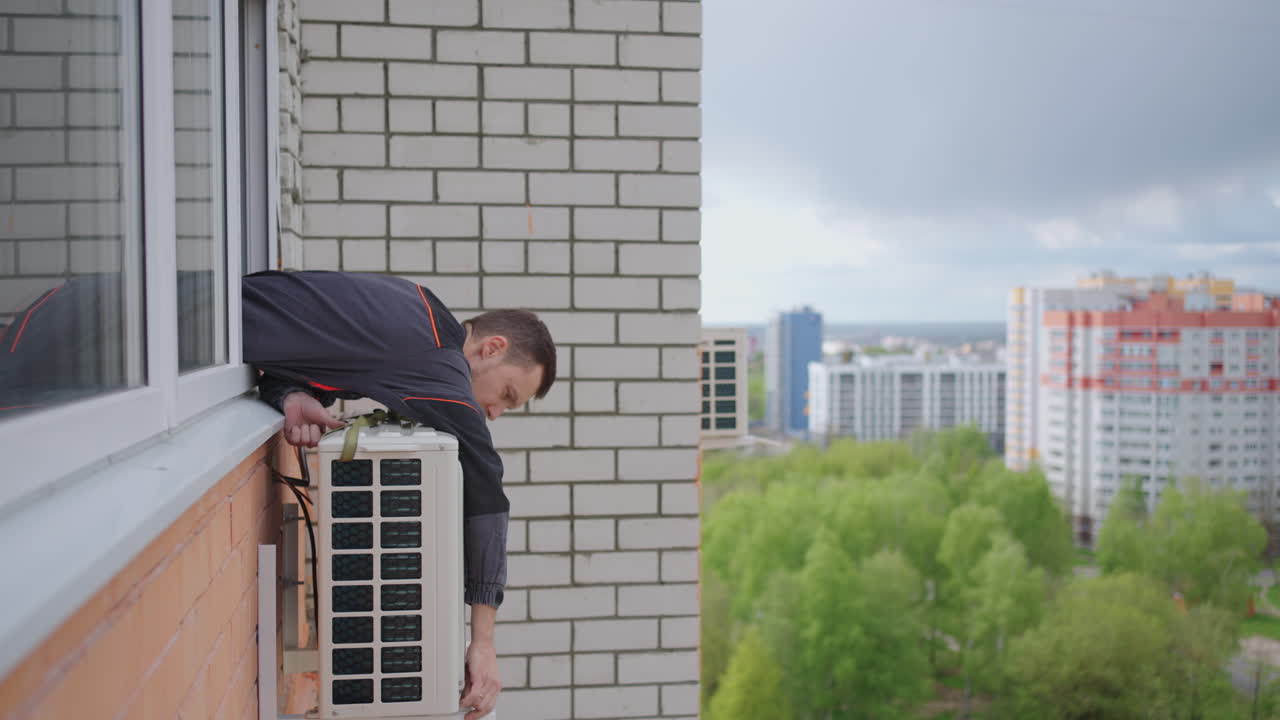 A man at high altitude installs air conditioning. Installation of the external unit of the Air Conditioning System. Creating a microclimate in the apartment