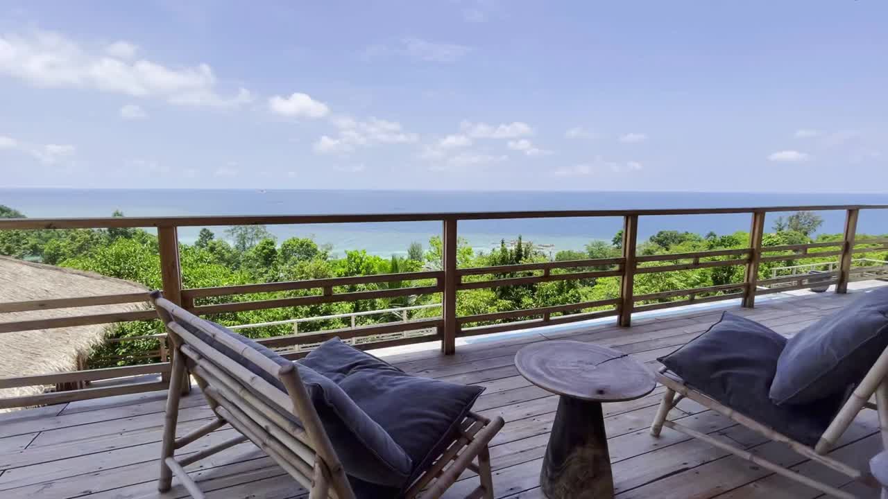 Tranquil deck view in Ko Samui with plush lounge chairs and table. Framed by lush greenery and ocean horizon. Bright, sunny, evokes relaxation and luxury