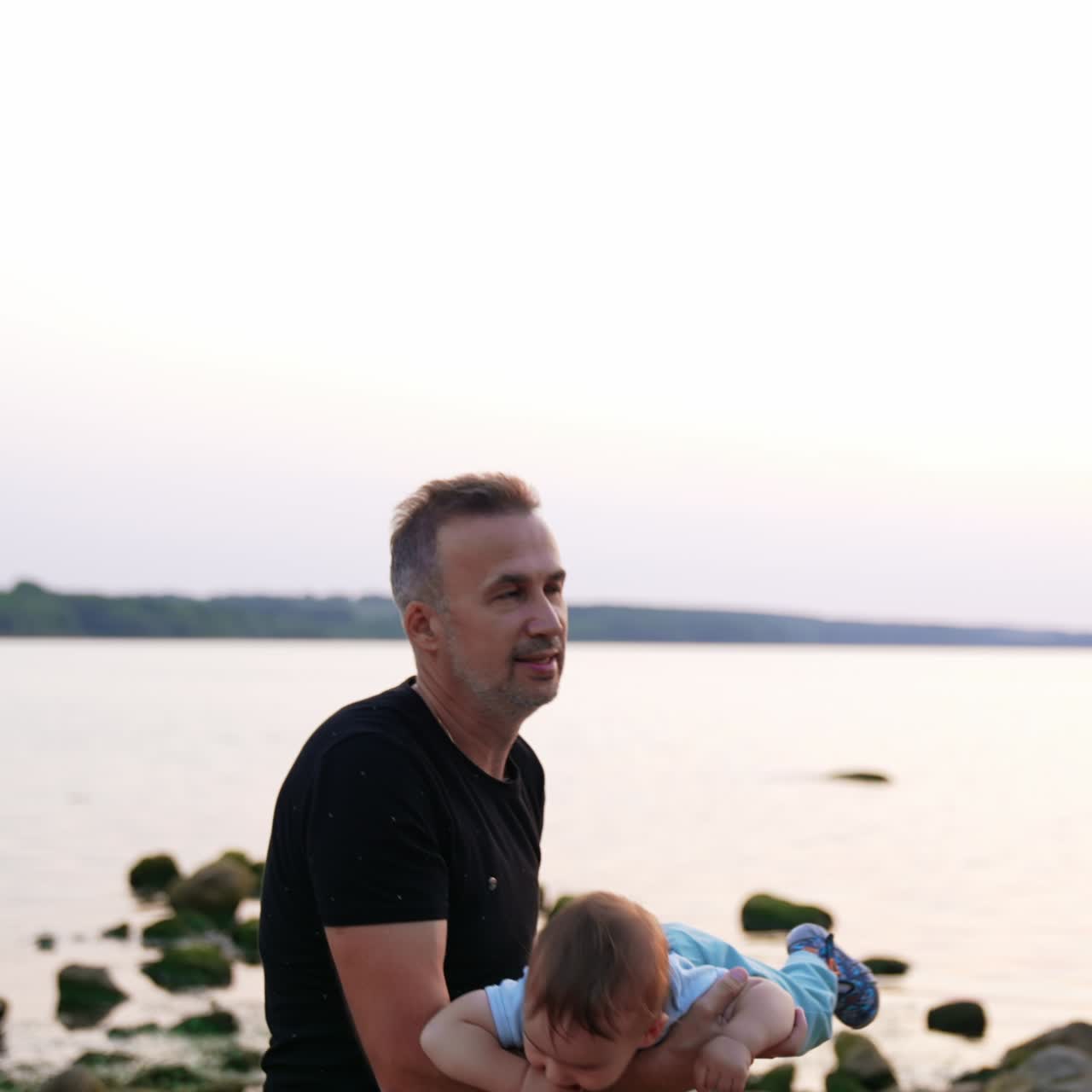Sweet little kid flies lying on his dad's arms. Adorable child smiling holding his finger in mouth. Lake at backdrop