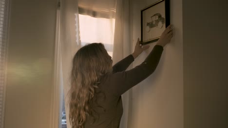 young blonde woman hanging up decor in her apartment
