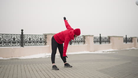 woman in red hoodie and gloves performing side stretches outdoors on snow-dusted path near decorative metal fence and lamp poles, surrounded by foggy atmosphere and serene urban winter landscape