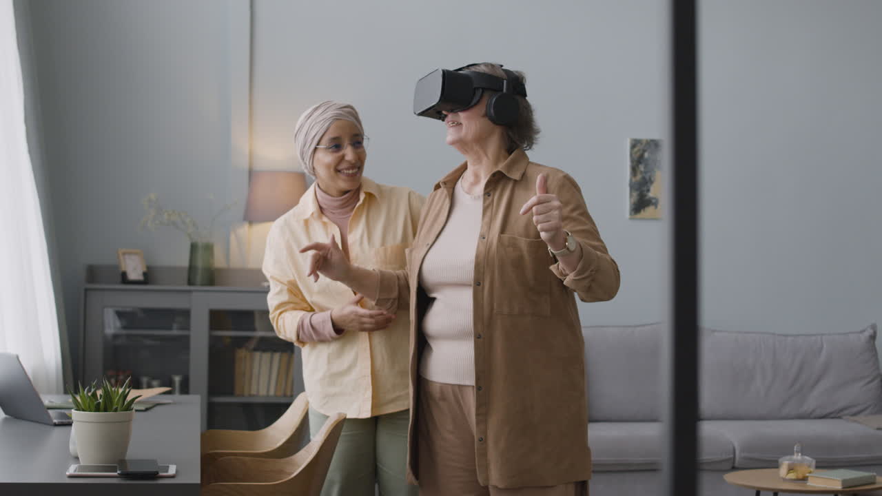 Middle Aged Arabic Woman Helping A Happy Senior Lady To Use Virtual Reality Headset Glasses At Home