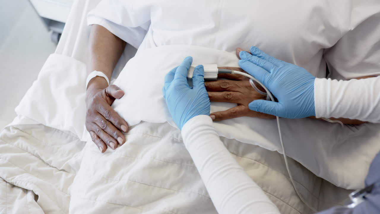 Hands of diverse female doctor and senior female patient in bed in hospital room, slow motion