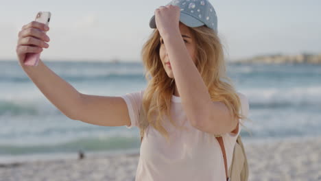 Retrato-De-Una-Mujer-Rubia-Feliz-Tomando-Una-Foto-Selfie-En-La-Playa-Usando-La-Tecnología-De-La-Cámara-De-Un-Teléfono-Inteligente-Disfrutando-Compartiendo-Experiencias-De-Vacaciones-Usando-Sombrero