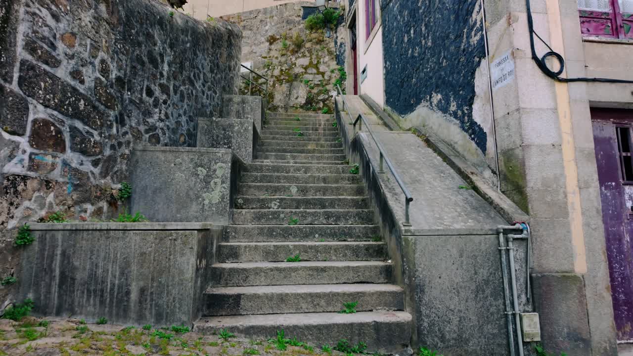 Escadaria de pedra e rampa paralela sobem entre edifícios antigos desgastados e muros de pedra rústicos em uma cidade histórica europeia. Apresenta uma porta roxa antiga distinta adicionando cor.