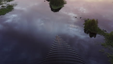 aerial drone shot of small motorboat with 2 passangers cruising slowly in a calm lake surface by golden hour sunset