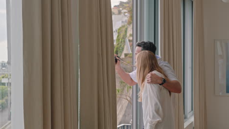 feliz pareja de viaje llegando a la habitación del hotel en vacaciones de luna de miel mirando por la ventana disfrutando de la hermosa vista del océano al atardecer abrazando compartir vacaciones románticas