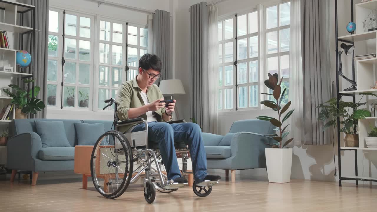 Asian Man Sitting In A Wheelchair While Use Mobile Phone Playing Video Game At Home