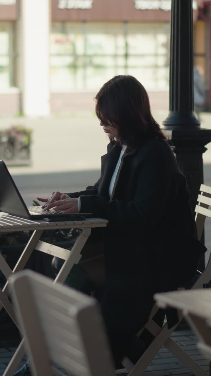 Student working on laptop outdoors at mall surrounded by wooden chairs, plants, and tripod, seated at table with coffee cup, paved walkway, bird flying down in the distance