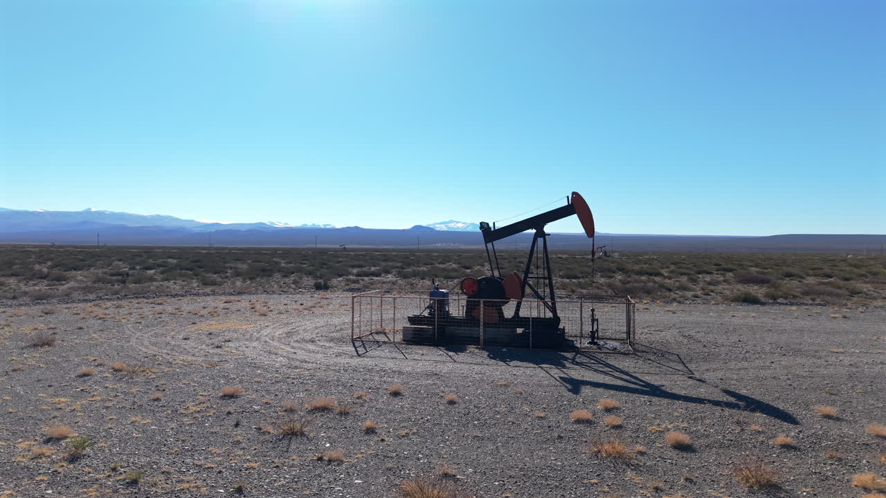 Drone captures oil pumpjack standing in a gravel desert plain near Malargüe, Mendoza, Argentina, with the Andes mountain range visible on the horizon under a clear sky, orbiting shot