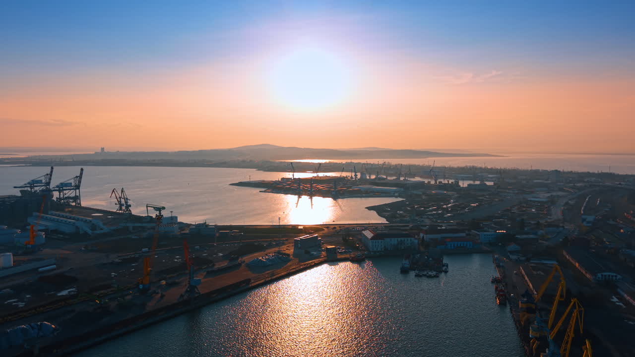 High angle view of maritime port under bright sun. Wide shot of harbor infrastructure, docks, and water reflection under a high sun. Clear sky and industrial zone