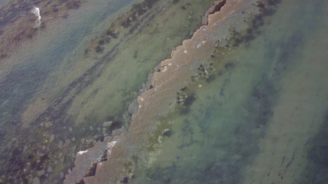 Drone video looking down over a ridged rock poking out of the rippling water. The footage moves to the right into Limmeridge Bay, Dorset. The last section over up over the cliffs surrounding the bay.