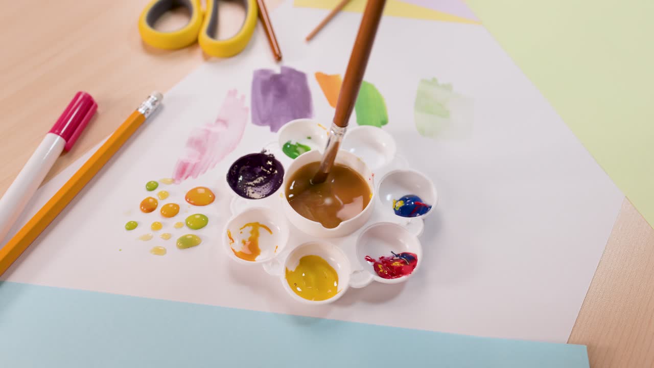 Child’s hand mixes paint with brush in palette, bright workspace, overhead view, natural lighting