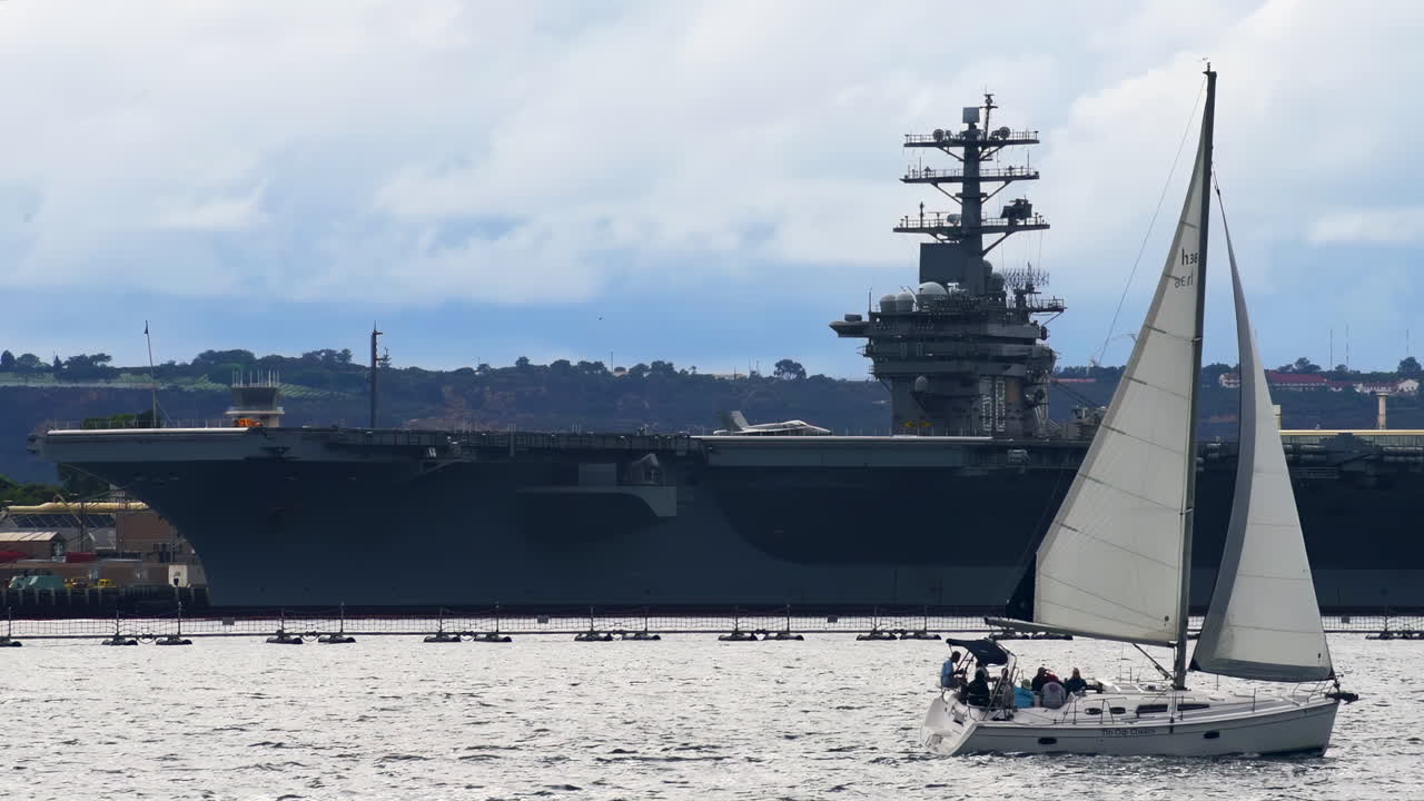 San Diego, California - September 26, 2021: Sailboat moving across San Diego Bay with a large U.S. Navy aircraft carrier in the background under cloudy sky
