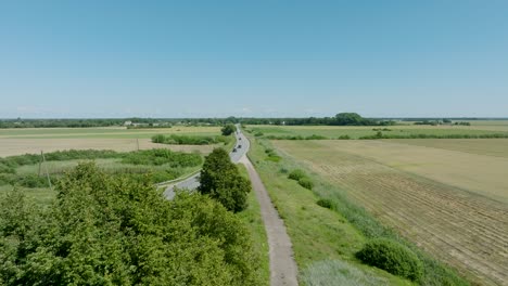 Aerial-establishing-shot-of-a-rural-landscape,-countryside-road-with-trucks-and-cars-moving,-lush-green-agricultural-crop-fields,-sunny-summer-day,-wide-drone-shot-moving-forward