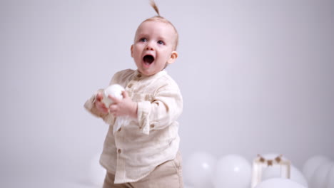 little caucasian baby stands up from the floor holding a white bunch in his hands. happy kid walks smiling adorably. white backdrop.