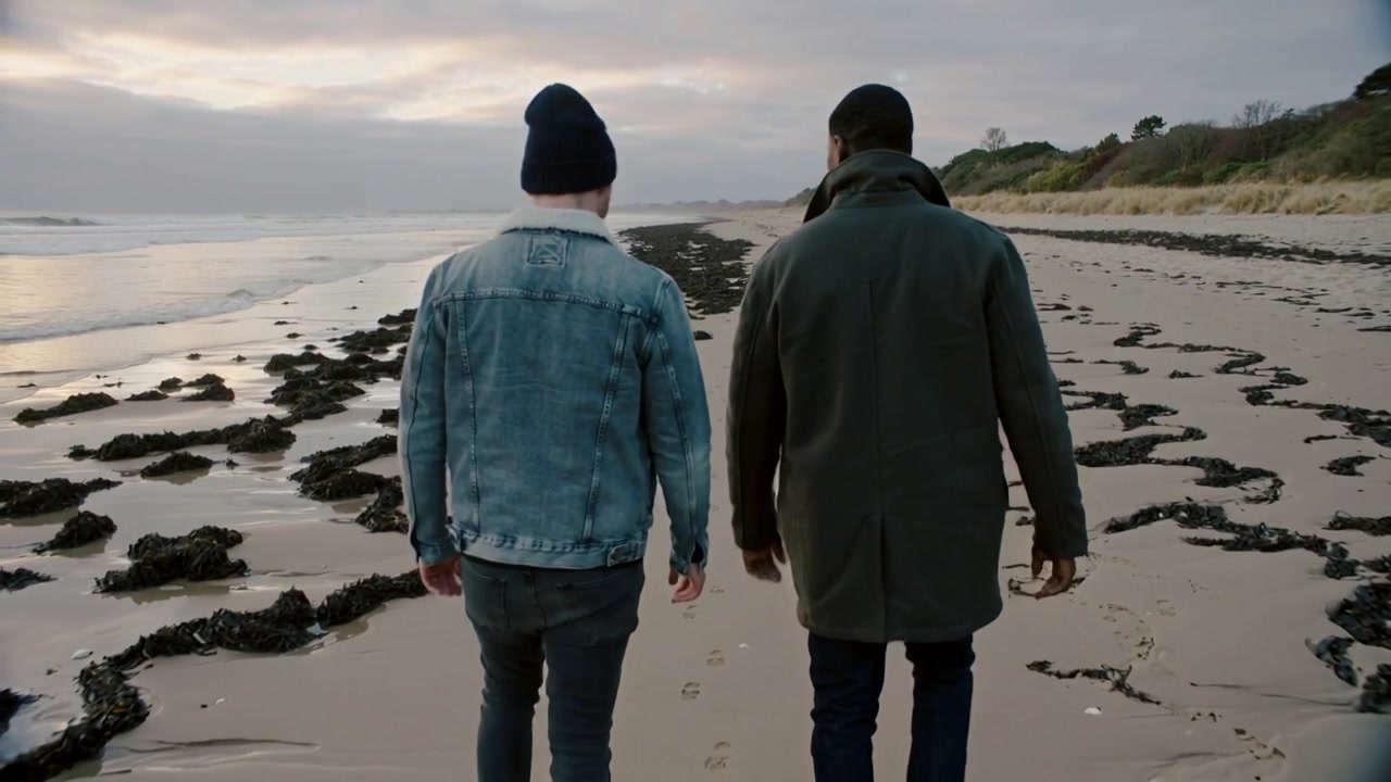 Walking men along shoreline after low tide exposes firm sand, talking, denim jacket beanie