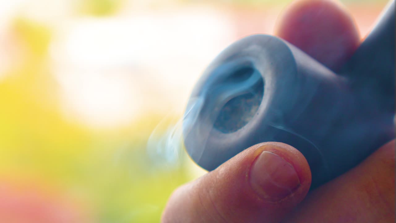 extreme closeup of a pure dark pipe with burned tobacco producing white blue smoke vintage antique style with a healthy fingers of a Caucasian male enjoying his moments sunny day blurry background