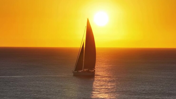 Um veleiro em silhueta move-se sobre águas calmas e abertas perto do horizonte, com um sol baixo e brilhante criando um brilho dourado quente e reflexos cintilantes no mar