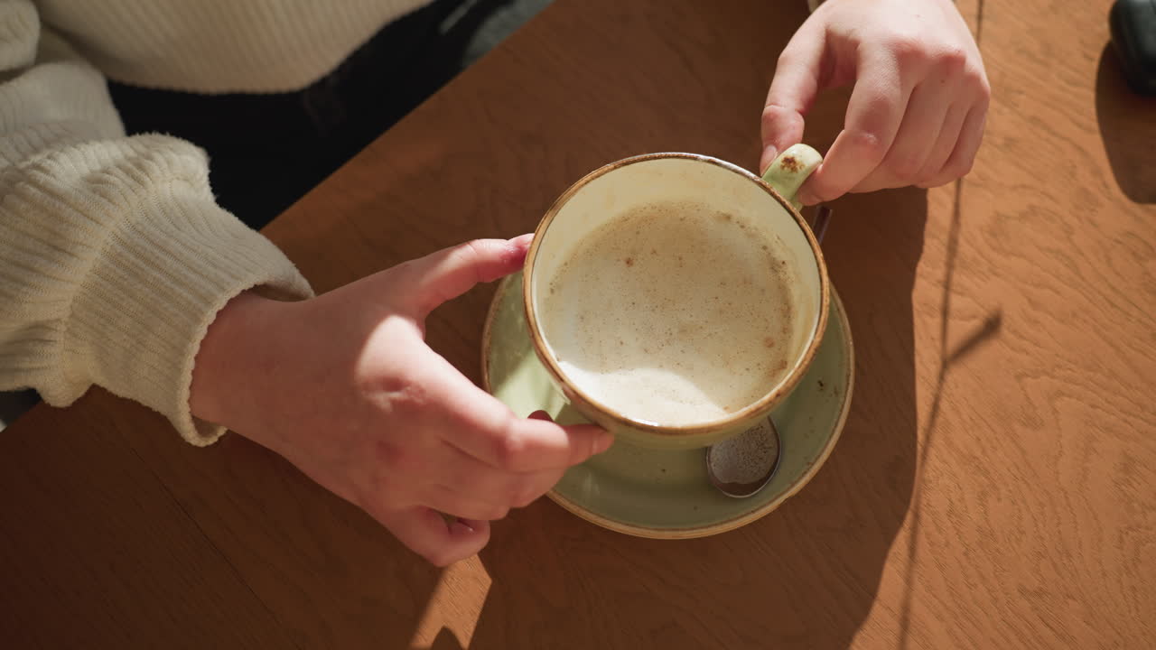 Aerial view of person in cozy sweater playfully rotating ceramic tea cup on wooden table, gentle morning sunlight casting soft shadows, relaxed indoor setting with spoon resting on saucer beside cup