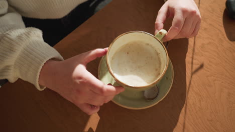 aerial view of person in cozy sweater playfully rotating ceramic tea cup on wooden table, gentle morning sunlight casting soft shadows, relaxed indoor setting with spoon resting on saucer beside cup