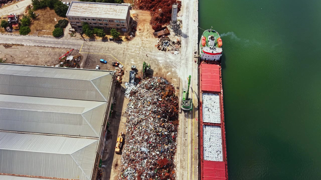 Heavy machinery moves scrap metal at a busy recycling center along the waterfront during daylight hours in a commercial area