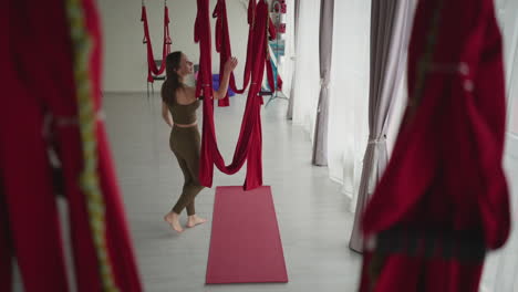 well lit yoga studio with floor to ceiling glass windows and urban house view as female athlete in sports wear swings gracefully on red silk hammock above mat, showing strength balance smooth motion