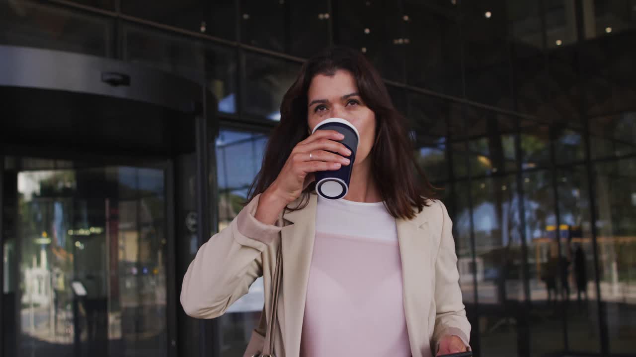 mujer de negocios caucásica usando un teléfono inteligente y bebiendo café para llevar fuera de una oficina moderna