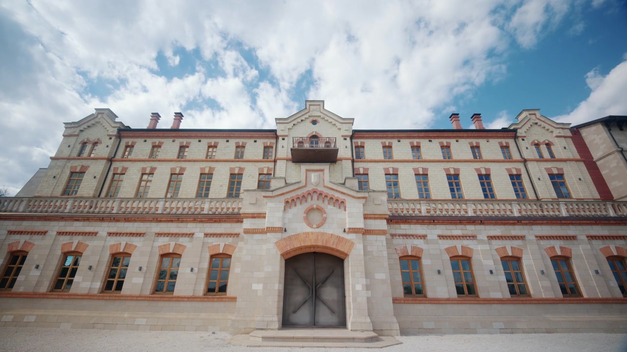 Široký čelní pohled na vinařský zámek v Moldavsku, s velkolepou historickou budovou, symetrickou fasádou, upravenými živými ploty, štěrkovou cestou a dramaticky modrou oblohou.