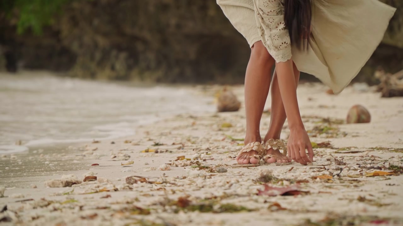 Donna a piedi nudi che cammina lungo una spiaggia sabbiosa, con le dita dei piedi che affondano nella sabbia bagnata, raccogliendo conchiglie e ciottoli, con un vestito estivo che svolazza, onde leggere che lambiscono le caviglie, legni levigati e rocce lisce sparsi, atmosfera calda.