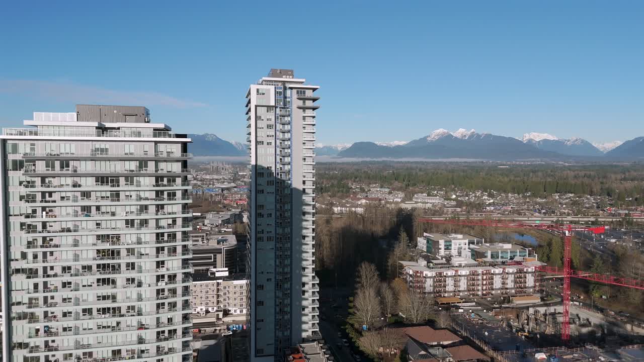 Arranha-céus modernos elevam-se sobre a cidade com áreas arborizadas e montanhas com picos nevados ao longe, em Langley, Colúmbia Britânica, Canadá - Tomada de drone em órbita.