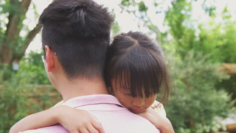 loving asian father cuddling daughter in garden as girl looks over his shoulder