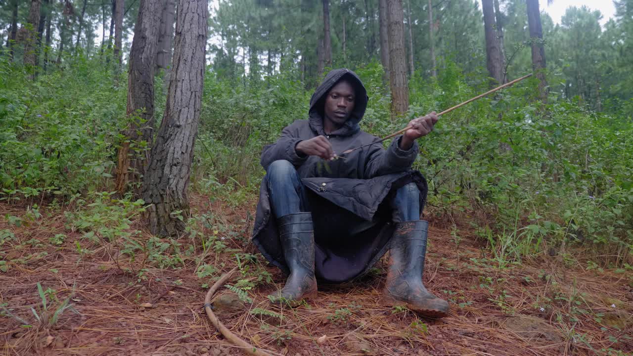 A African hunter wearing a hooded cloak and gum boots sits down on the forest floor and cleans his bow and arrow with leaves