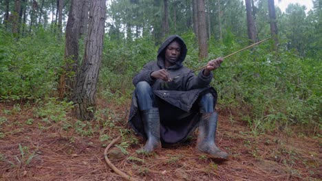 a african hunter wearing a hooded cloak and gum boots sits down on the forest floor and cleans his bow and arrow with leaves