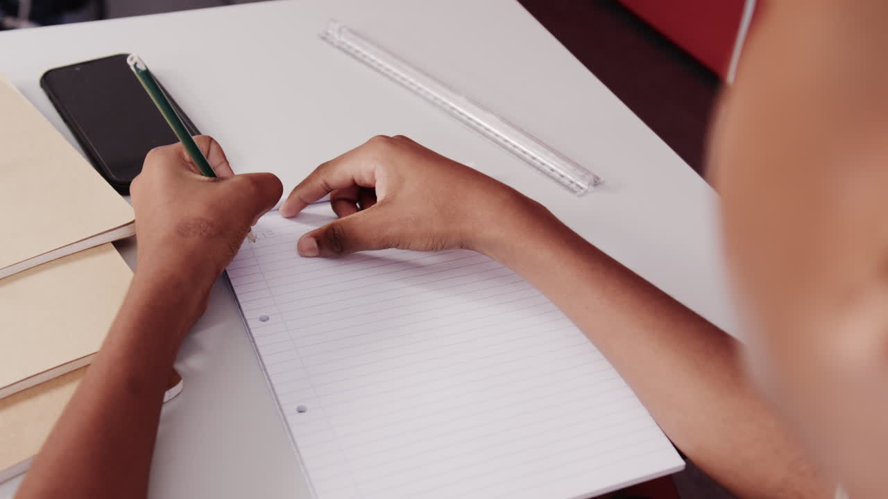 Writing in notebook, student using pencil and smartphone at school desk