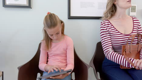 Caucasian-girl-patient-using-tablet-sitting-with-mother-in-hospital-reception,-slow-motion