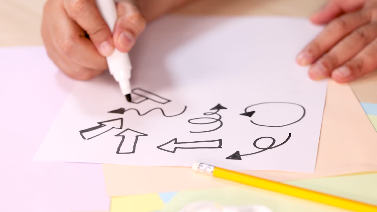 Person draws various black arrows and doodles on white paper using marker, top-down view