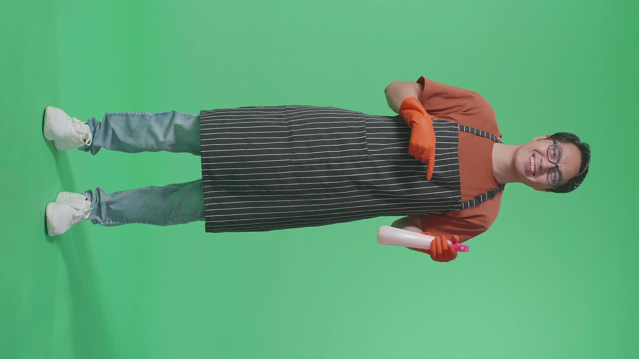 Full Body Of Smiling Asian Male Housekeeper With An Apron Pointing A Spray While Standing In The Green Screen Studio
