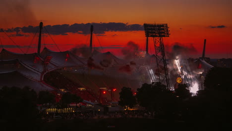 Rammstein-Till-Lindemann-German-metal-band-concert-show-with-pyrotechnics,-fire,-smoke,-flames-in-the-famous-Germany-Munich-Olympic-Stadium-by-evening-sunset-sky-and-a-huge-crowd-of-fans