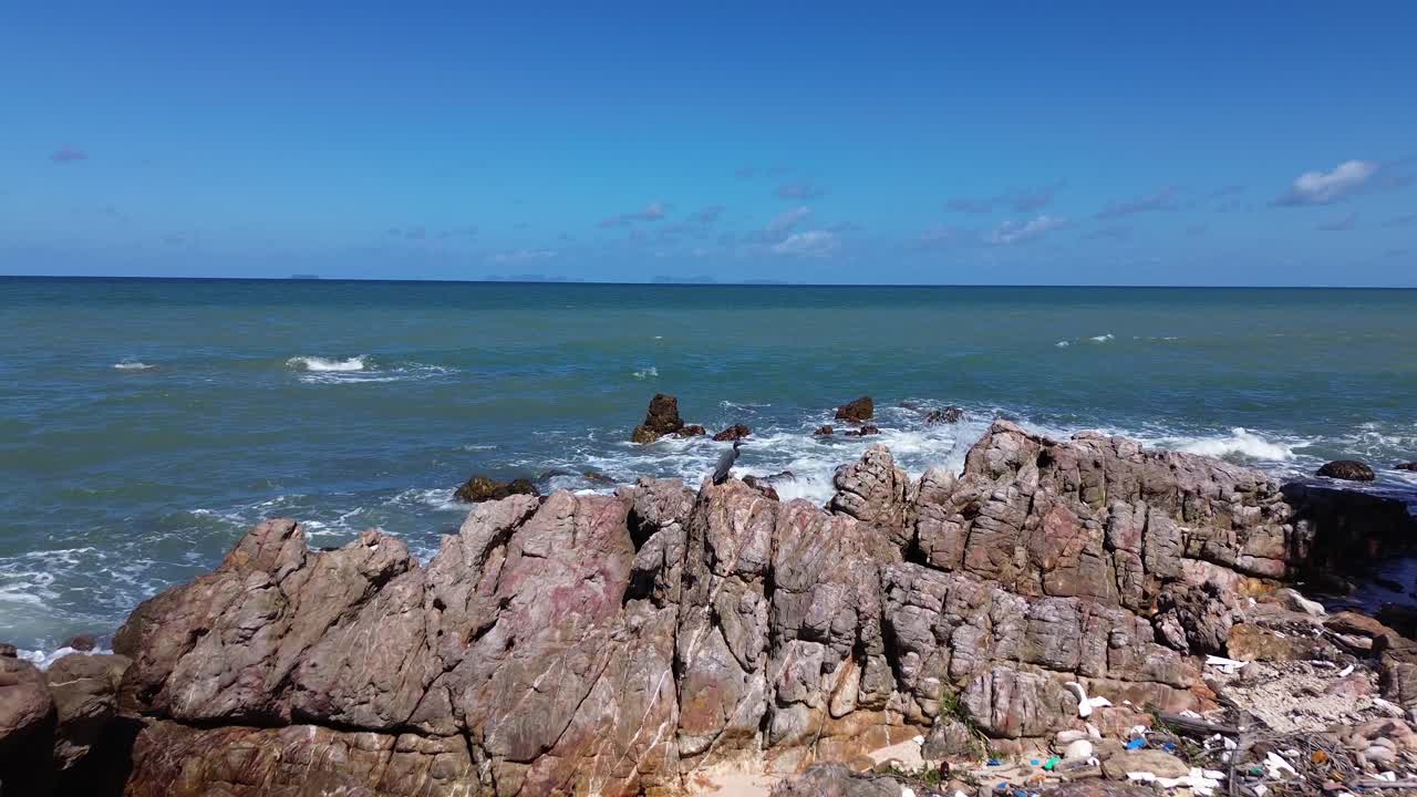 Aerial drone footage of a bird perched on rocky shoreline with waves crashing on the coast in Thailand, scenic seascape with wildlife and rugged tropical environment