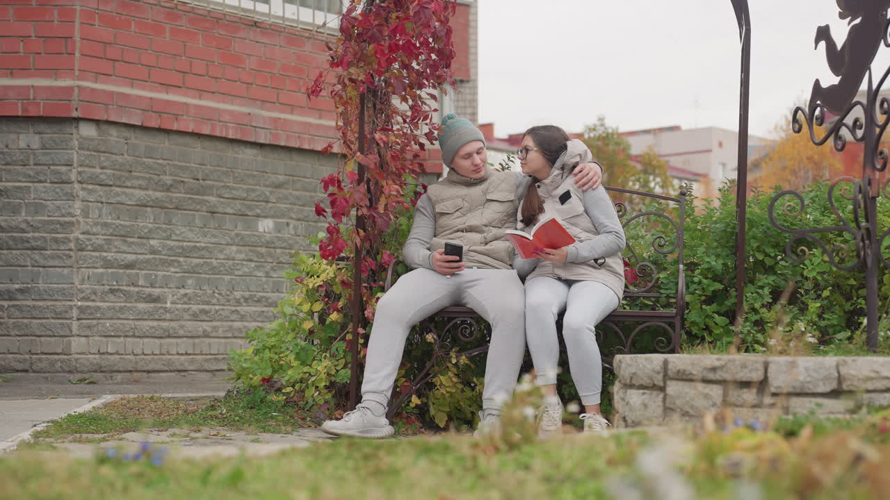 Lovers seated outdoors on garden bench in cool breeze as man warmly pulls woman close while she reads novel, surrounded by greenery, flowers, and brick walls in cozy autumn urban setting