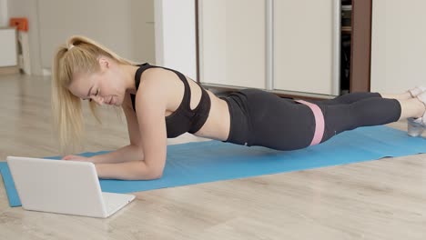 chica sexy rubia haciendo su entrenamiento de gimnasio en casa mientras ve lecciones de video en línea en la computadora portátil