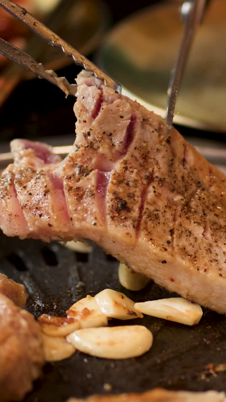 Primo piano verticale di mani che usano forbici e pinze per tagliare carne di maiale su una griglia.