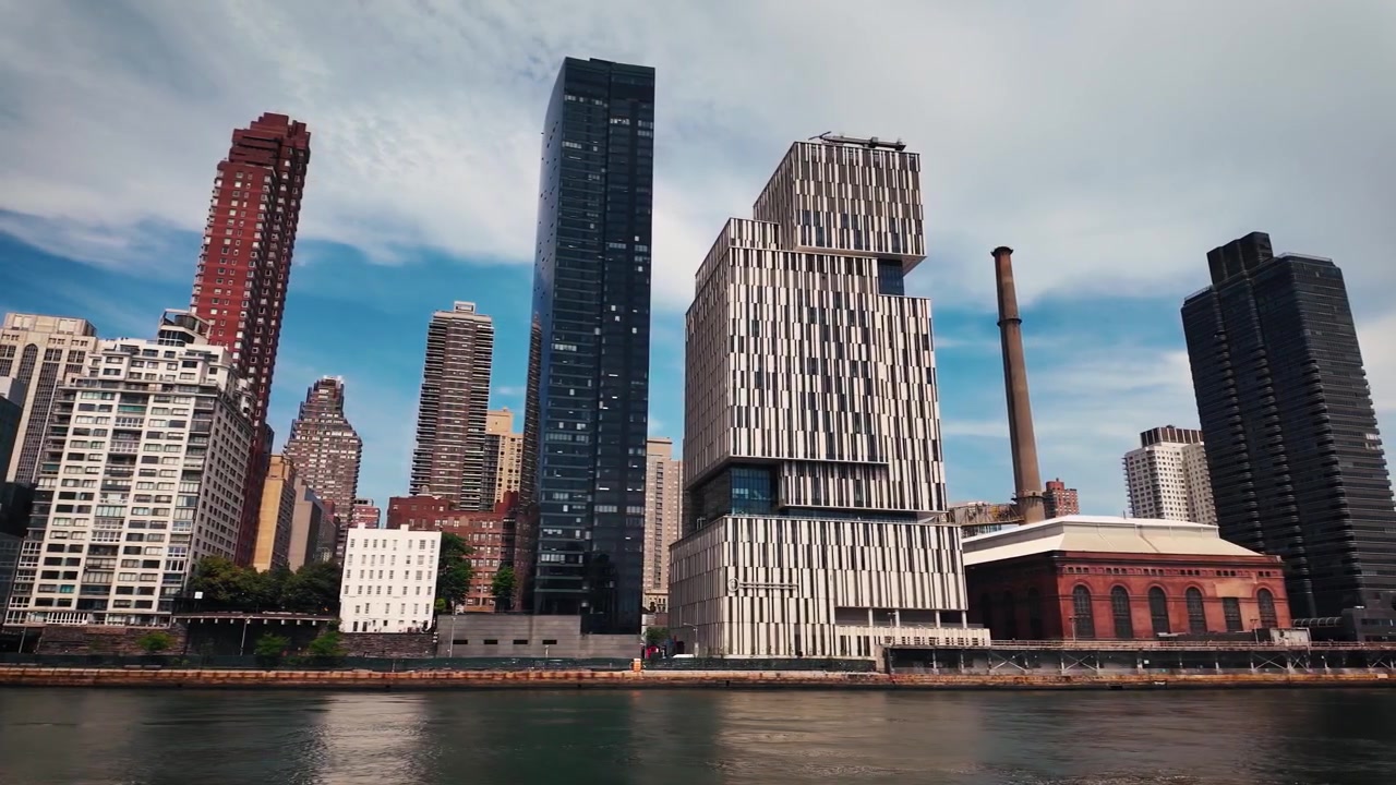 Una barca scivola lungo il lungomare dell'East River di Roosevelt Island, caratterizzato dal campus Cornell Tech, torri residenziali e una storica centrale elettrica sotto nuvole drammatiche