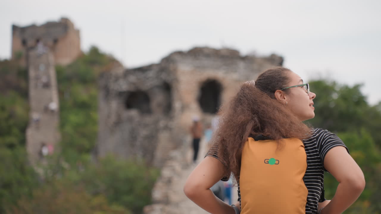 Traveler ransel mengagumi reruntuhan batu kuno, tampak belakang penjelajah muda dengan ransel oranye dan rambut keriting, tangan di pinggul sambil memindai menara yang hancur dan lereng bukit, suasana kontemplatif dengan nuansa budaya.