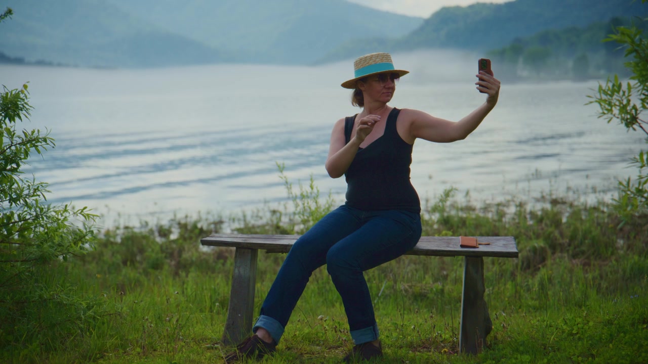 Eine Frau sitzt auf einer Holzbank an einem ruhigen See. Sie trägt einen Hut und hält ein Handy, um Selfies zu machen. Nebel steigt aus dem Wasser auf und umhüllt sanft die fernen Berge. Grünes Gras umgibt sie.