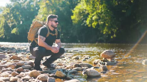 Male-hiker-crouching-by-a-river-surounded-by-rocks-and-trees,-looking-around-during-hike-in-jungle